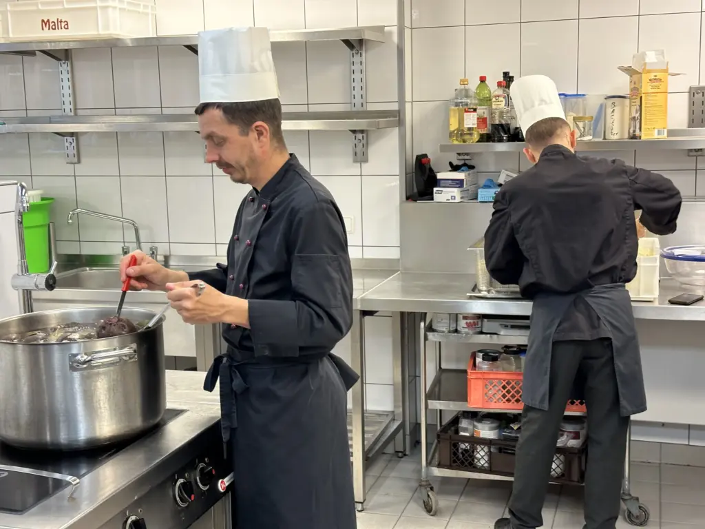 Zwei Köche im Haus Tannenberg Gummersbach bei der Arbeit in der Küche – einer rührt in einem großen Topf, der andere bereitet Zutaten auf der Arbeitsfläche vor.